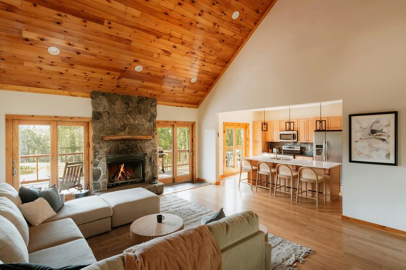 Open living room with a stone fireplace, wood ceiling, and kitchen beyond